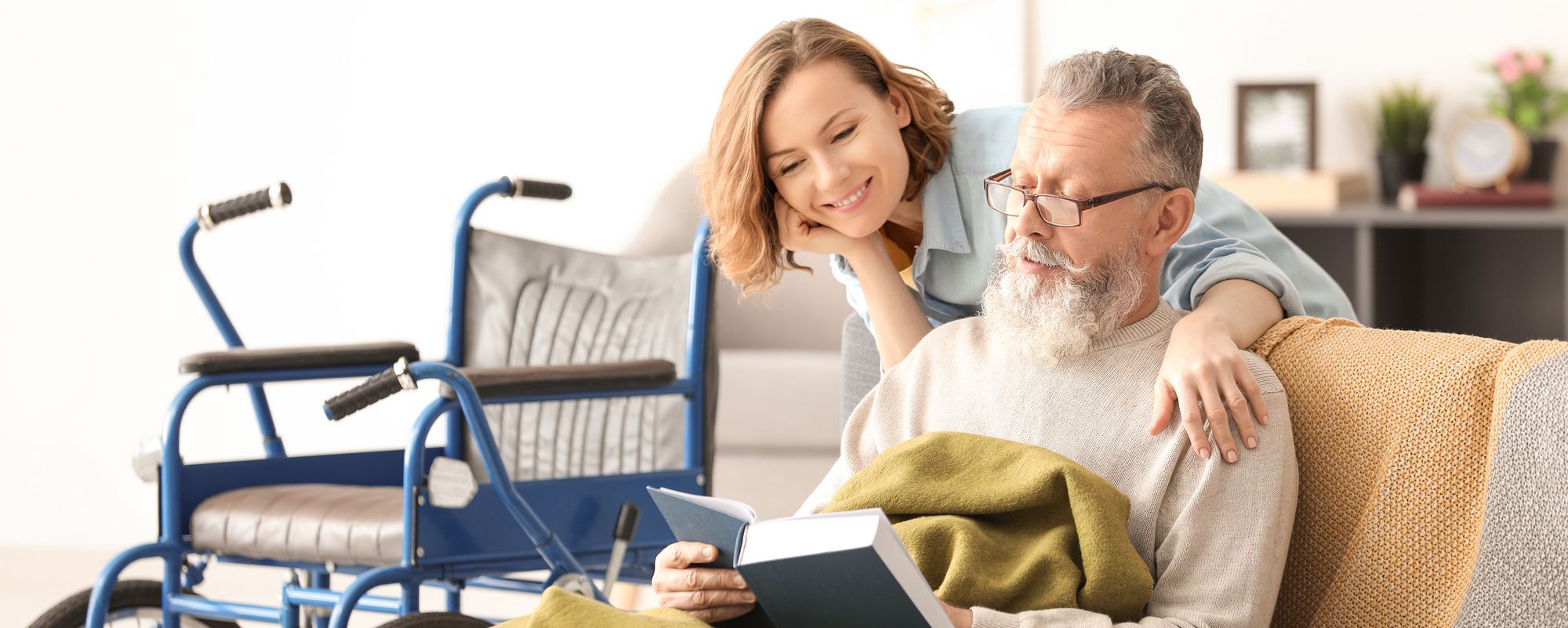 Pflegekraft liest älterem Mann im Rollstuhl zu Hause vor